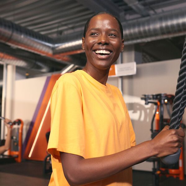 Una mujer sonr&iacute;e mientras sostiene una battle rope durante un entrenamiento en un gimnasio Basic-Fit. Equipos de fuerza y detalles en color naranja son visibles al fondo.