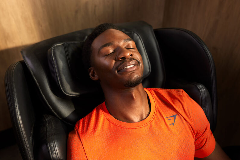 Un hombre se relaja en una silla de masaje en un gimnasio Basic-Fit, con los ojos cerrados y sonriendo despu&eacute;s de su entrenamiento. Lleva una camiseta deportiva naranja en una zona de recuperaci&oacute;n tranquila.