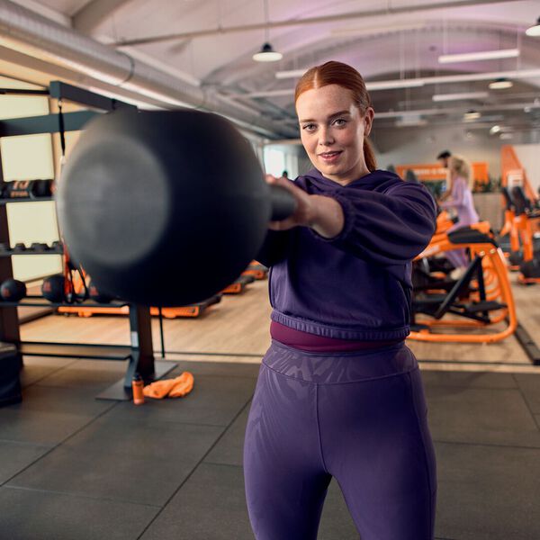 Una mujer realiza un kettlebell swing durante un entrenamiento en un gimnasio Basic-Fit, entrenando en la zona funcional con equipamiento visible al fondo.