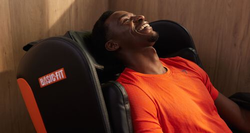 Een man in een oranje sportshirt ontspant en glimlacht terwijl hij in een zwarte Basic-Fit massagestoel zit.