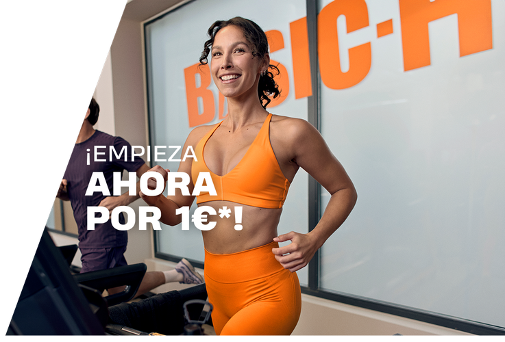 A woman performs a barbell back squat in a Basic-Fit gym, smiling while lowering into the squat with a weighted bar on her shoulders. The gym equipment and warm lighting create an active training atmosphere.