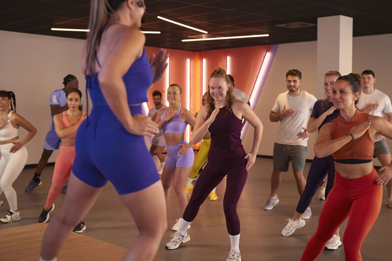 Groep mensen die meedoet aan een Shape-les in een fitnessstudio, met een instructeur vooraan.