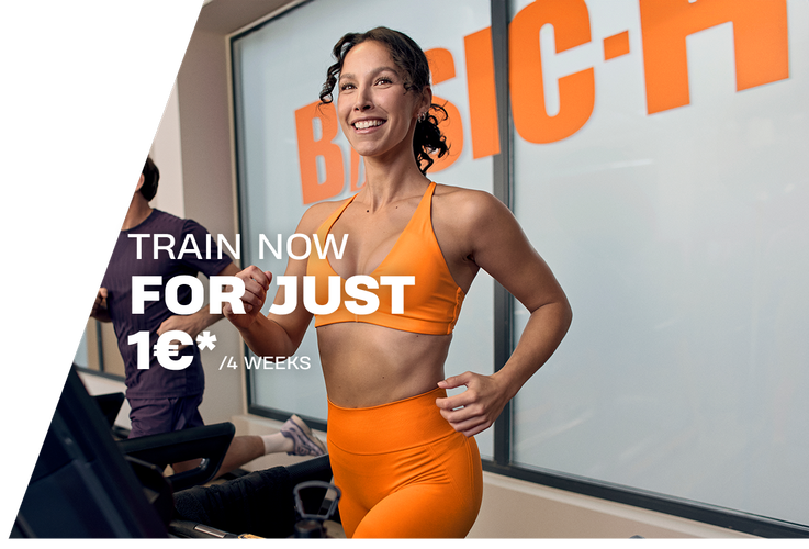 A woman performs a barbell back squat in a Basic-Fit gym, smiling while lowering into the squat with a weighted bar on her shoulders. The gym equipment and warm lighting create an active training atmosphere.