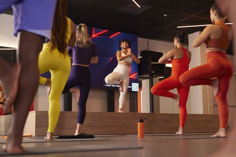 Yogainstructeur die een groepsles leidt vanaf een podium terwijl deelnemers balansoefeningen op &eacute;&eacute;n been doen op hun matten