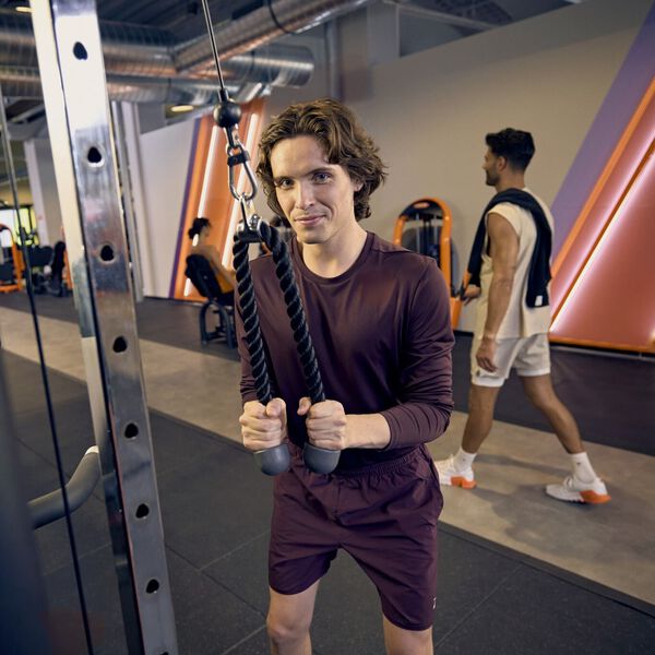 Un hombre realiza un pushdown de tr&iacute;ceps con cuerda en una m&aacute;quina de poleas en un gimnasio Basic-Fit, concentrado en su entrenamiento mientras otros miembros entrenan al fondo.