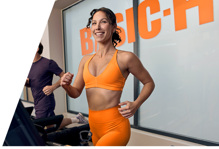 A woman performs a barbell back squat in a Basic-Fit gym, smiling while lowering into the squat with a weighted bar on her shoulders. The gym equipment and warm lighting create an active training atmosphere.