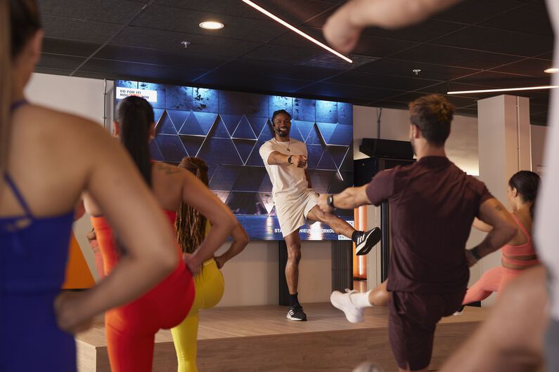 Groepsfitnessles met een instructeur op een podium die een beweging toont, terwijl deelnemers op de vloer meedoen.