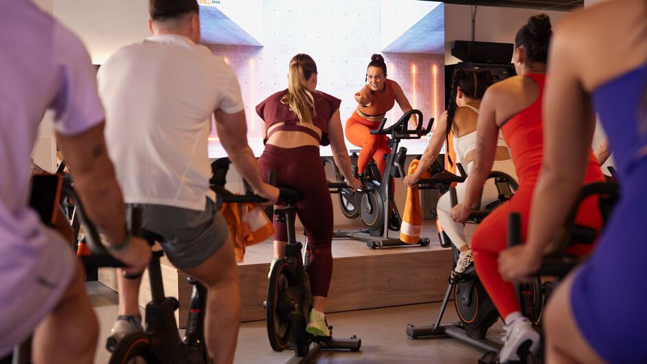 A group cycles on stationary bikes while an instructor on a raised platform leans forward on the handlebars and looks toward the participants.