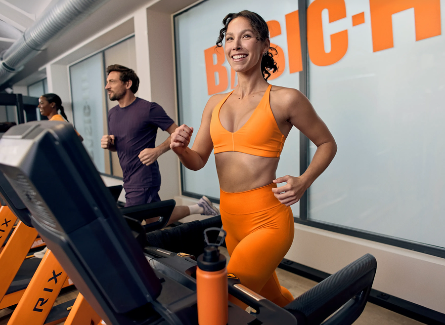 A woman performs a barbell back squat in a Basic-Fit gym, smiling while lowering into the squat with a weighted bar on her shoulders. The gym equipment and warm lighting create an active training atmosphere.