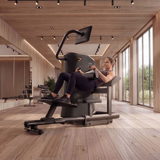 Frau nutzt eine sitzende EGYM-Beinpresse in einem hellen Fitnessstudio mit Spiegeln, Holzw&auml;nden und gro&szlig;en Fenstern.