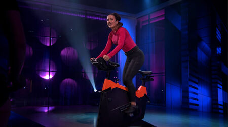 Woman cycling on an indoor bike under dramatic purple lighting in a dark studio.	