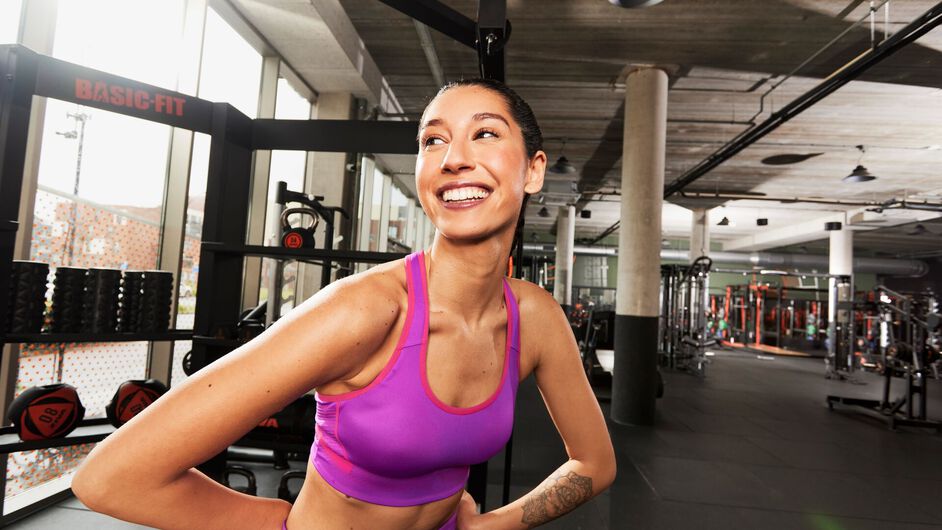 Woman in a pink and purple sports bra smiling confidently in a Basic-Fit club, standing with hands on hips post-workout.