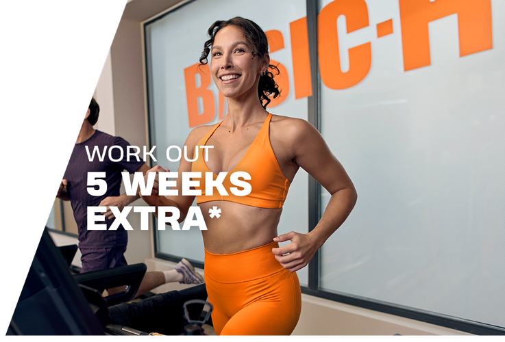 A woman performs a barbell back squat in a Basic-Fit gym, smiling while lowering into the squat with a weighted bar on her shoulders. The gym equipment and warm lighting create an active training atmosphere.