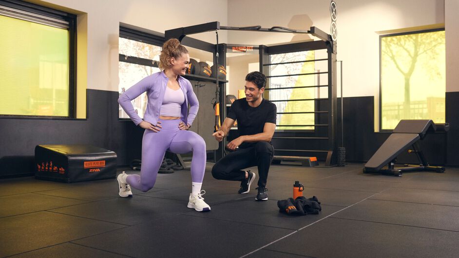 Personal trainer begeleidt een vrouw tijdens een lunge oefening in een functionele trainingsruimte in een Basic-Fit gym