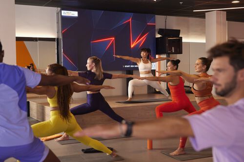 Groep sporters die deelnemen aan een yogaachtige groepsles in een fitnessstudio, in een brede staande houding met gestrekte armen terwijl een instructeur lesgeeft vanaf een podium.