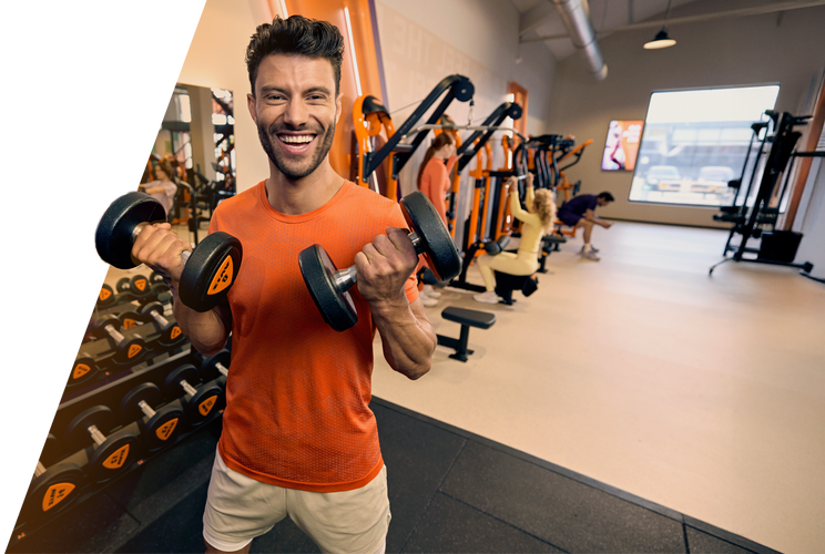 Ein Mann l&auml;chelt, w&auml;hrend er bei einem Krafttraining im Basic-Fit Fitnessstudio Kurzhanteln hebt, w&auml;hrend andere Mitglieder im Hintergrund trainieren.