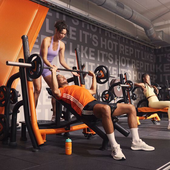 Een sporter voert een incline bench press uit met ondersteuning van een trainer bij Basic-Fit. Op de achtergrond trainen andere leden op krachttoestellen.
