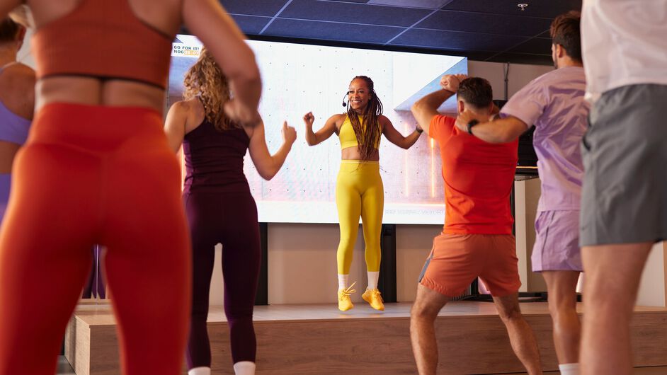 Los participantes participan en una clase de fat burn cardio y realizan movimientos continuos juntos siguiendo al instructor al frente de la sala.