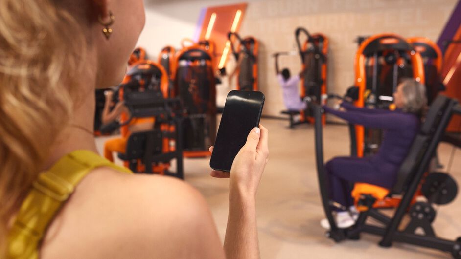 Persoon gebruikt de Basic-Fit app op de telefoon om een groepsles te boeken in de club terwijl anderen op toestellen trainen.