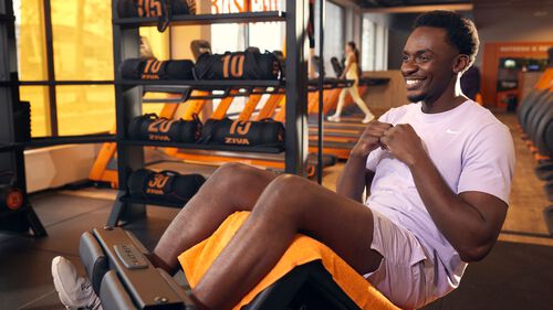 Man doing seated ab crunches on a gym machine, smiling during his workout with sandbags and cardio equipment in the background