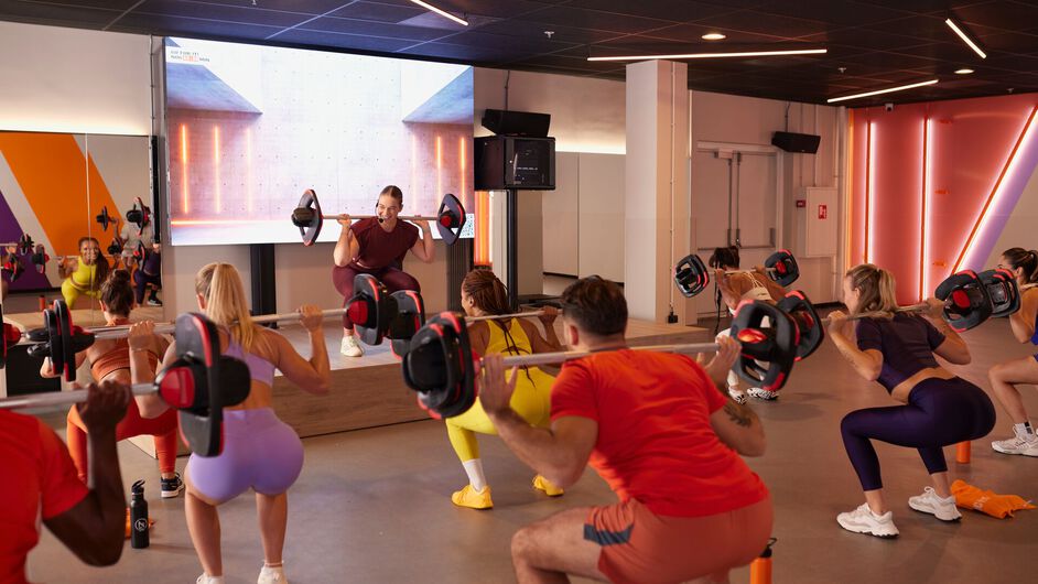 A group follows a high-energy workout, raising their arms and moving in sync while an instructor leads from a stage in a fitness studio.