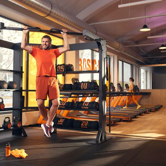 Een sporter doet een pull-up aan een functioneel trainingsrek bij Basic-Fit, terwijl een ander lid op een loopband loopt.