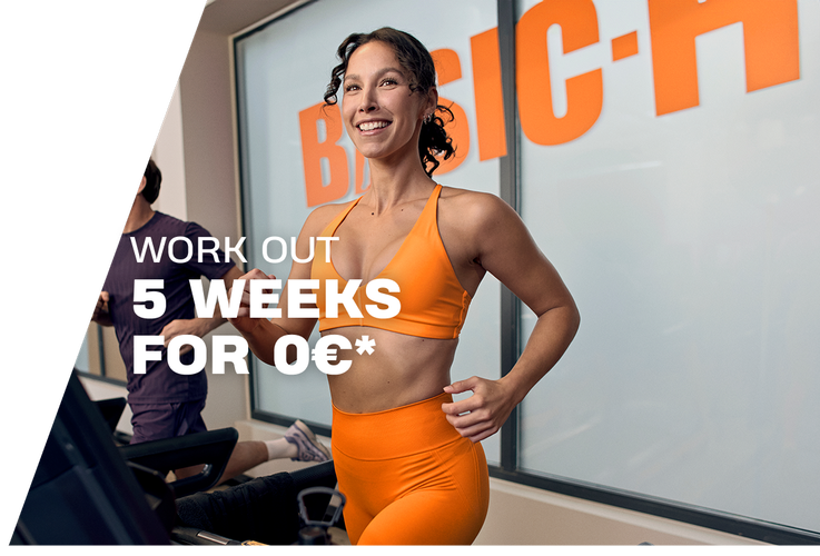 A woman performs a barbell back squat in a Basic-Fit gym, smiling while lowering into the squat with a weighted bar on her shoulders. The gym equipment and warm lighting create an active training atmosphere.