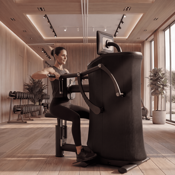 Frau nutzt eine sitzende EGYM-Brustpresse in einem hellen Fitnessstudio mit Hanteln und gro&szlig;en Fenstern.