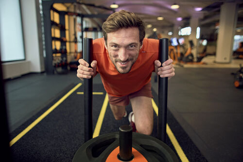 Un hombre empuja un trineo con peso durante un entrenamiento en un gimnasio Basic-Fit y sonr&iacute;e mientras entrena en la zona de entrenamiento funcional.