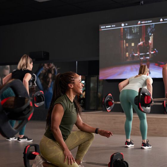 Een groep leden volgt een virtuele krachttraining via het GXR-scherm bij Basic-Fit en voert samen oefeningen met een halterstang uit.