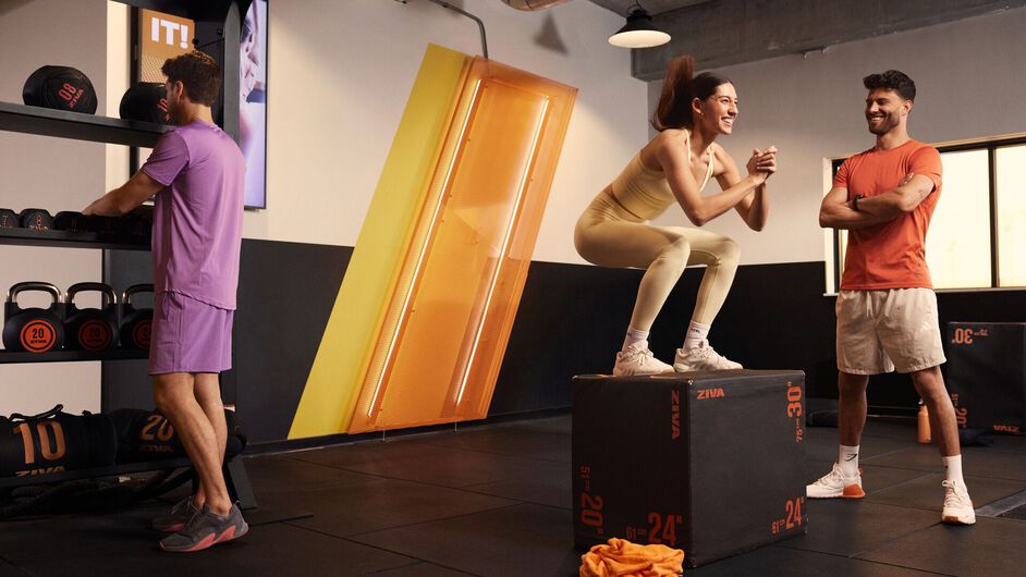 A group follows a high-energy workout, raising their arms and moving in sync while an instructor leads from a stage in a fitness studio.