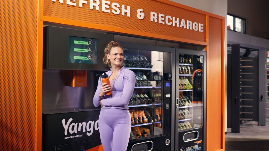 Una mujer est&aacute; de pie con su botella en una estaci&oacute;n de YANGA Sports Water en un gimnasio Basic-Fit, sonriendo.
