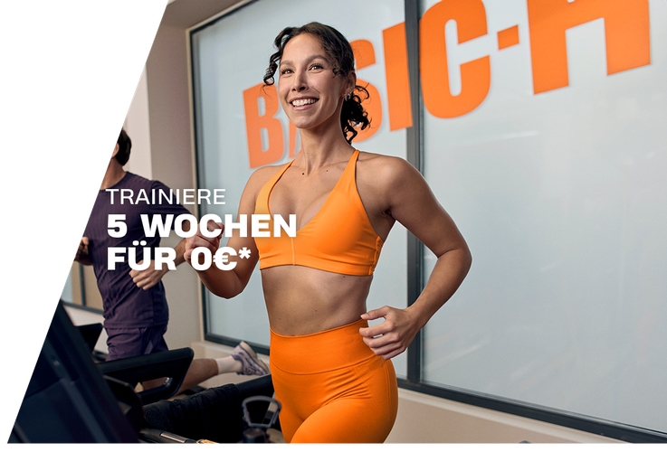 A woman performs a barbell back squat in a Basic-Fit gym, smiling while lowering into the squat with a weighted bar on her shoulders. The gym equipment and warm lighting create an active training atmosphere.