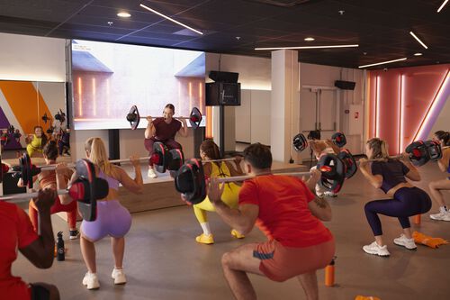A group of people performs squats with barbells while an instructor demonstrates the movement on a raised stage in a fitness studio.