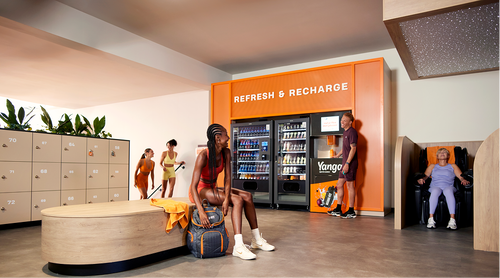 Members relaxing and chatting in a Basic-Fit lounge with lockers, seating and a refreshment vending area.