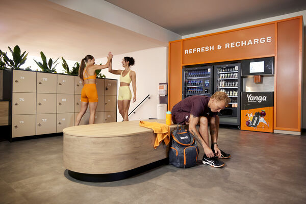 Mitglieder bereiten sich im Umkleidebereich eines Basic-Fit Clubs auf ihr Training vor, w&auml;hrend sich zwei Sportler begr&uuml;&szlig;en.