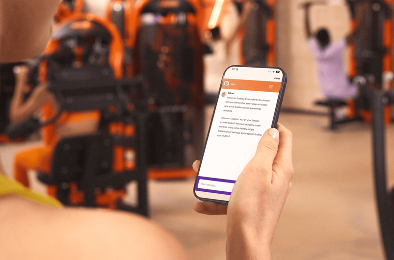 Persoon houdt een smartphone vast in een Basic-Fit gym en gebruikt de app voor workout begeleiding terwijl fitnessapparatuur op de achtergrond zichtbaar is