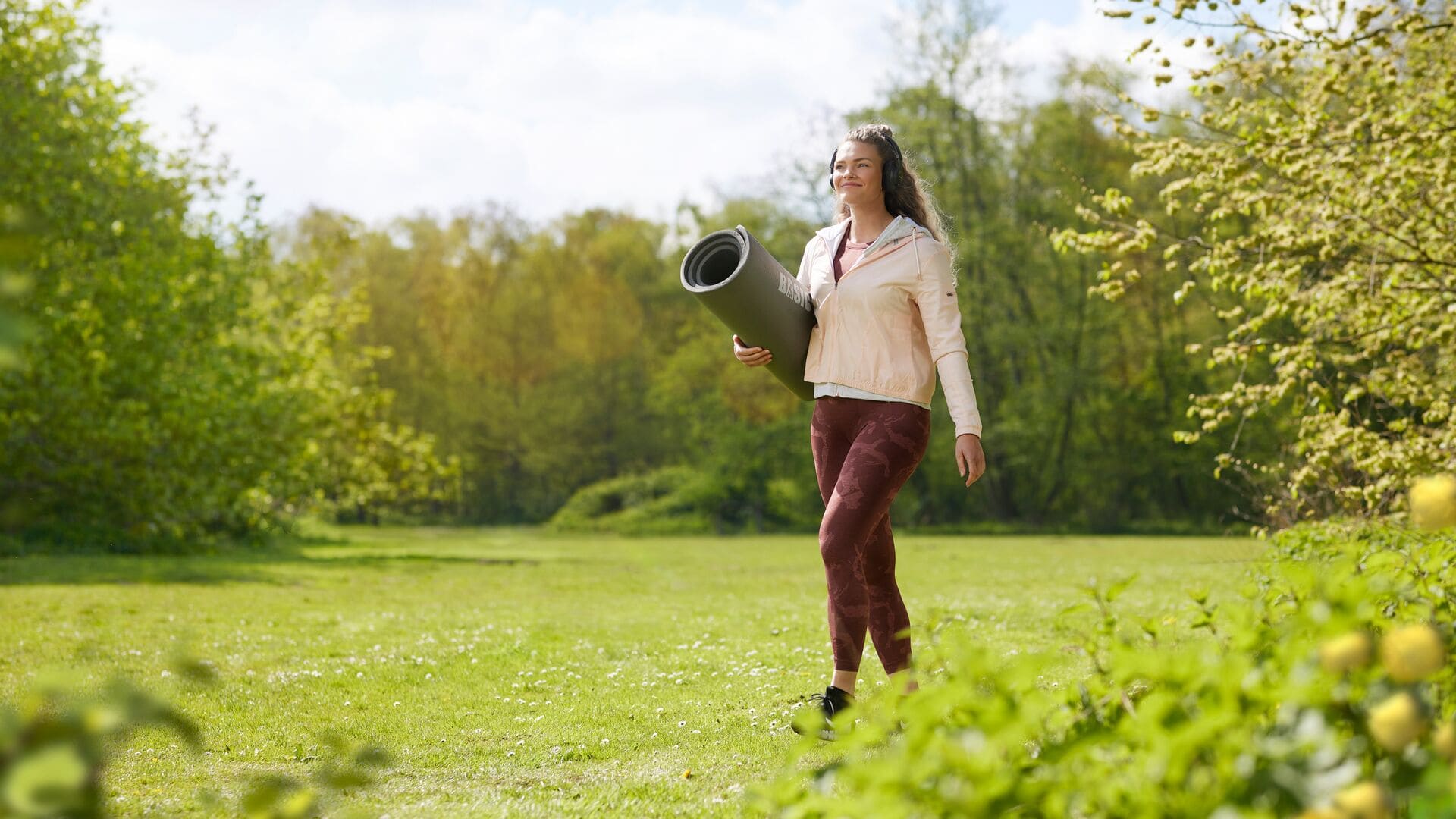 Une femme marche dans un parc verdoyant avec un tapis de yoga enroulé sous le bras, souriante et habillée en tenue de sport.