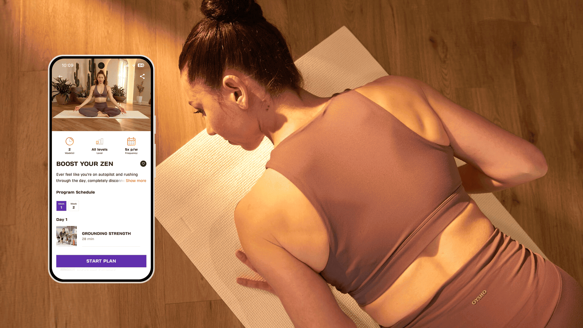 A woman lies on a yoga mat in a softly lit room, preparing for a calm workout. She wears mauve activewear and focuses on her breath. Next to her, a smartphone shows the Basic-Fit program “Boost Your Zen,” offering sessions to restore balance and inner strength.