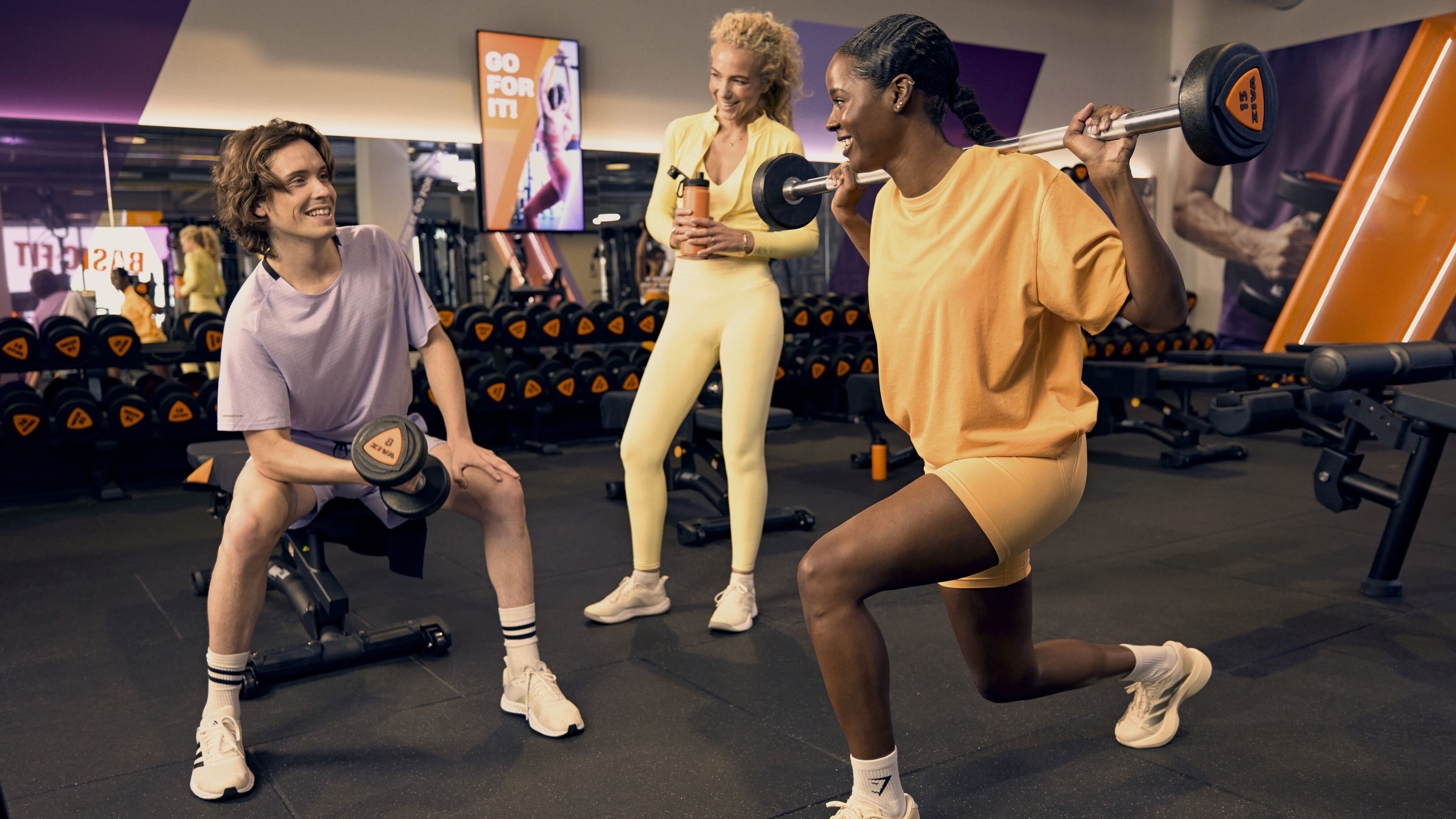 Tres personas entrenan juntas en la zona de peso libre, una hace una zancada con barra mientras las otras animan y sonríen