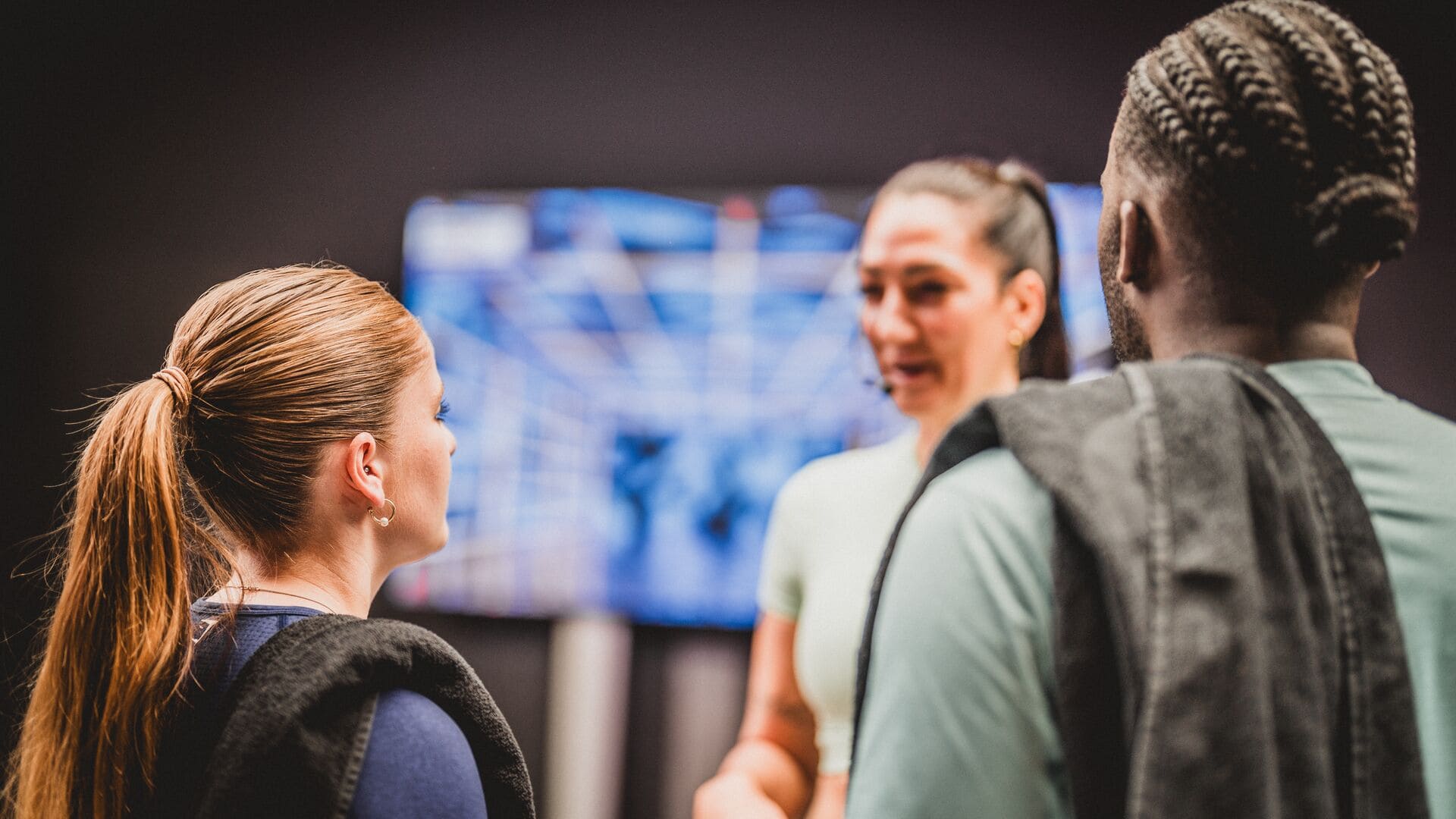 Two Basic-Fit members listen attentively to a trainer during an introduction or class briefing, with a screen displaying gym visuals in the background.        Ask ChatGPT