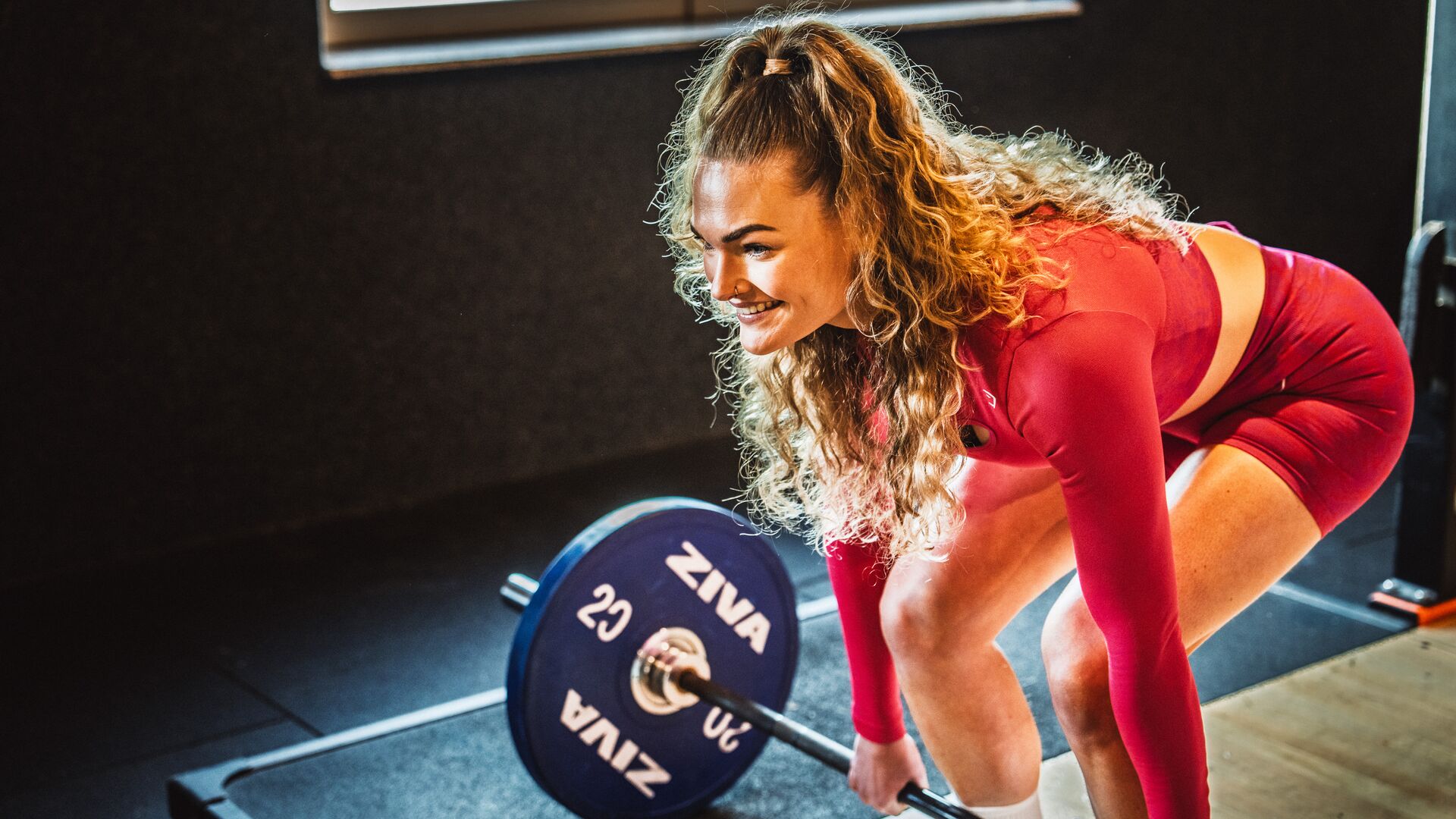 A woman in a red outfit preparing to lift a barbell with blue weight plates in a gym. She is smiling and focused on her workout.