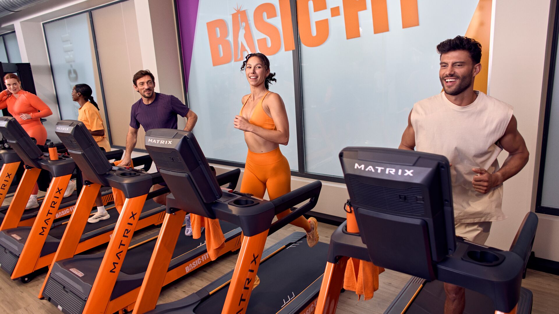 Eine Gruppe von Trainierenden läuft auf orangefarbenen Matrix-Laufbändern in einem hellen Basic-Fit-Studio, mit einer Frau in orangefarbener Sportkleidung und einem lächelnden Mann in einem ärmellosen Oberteil im Vordergrund.