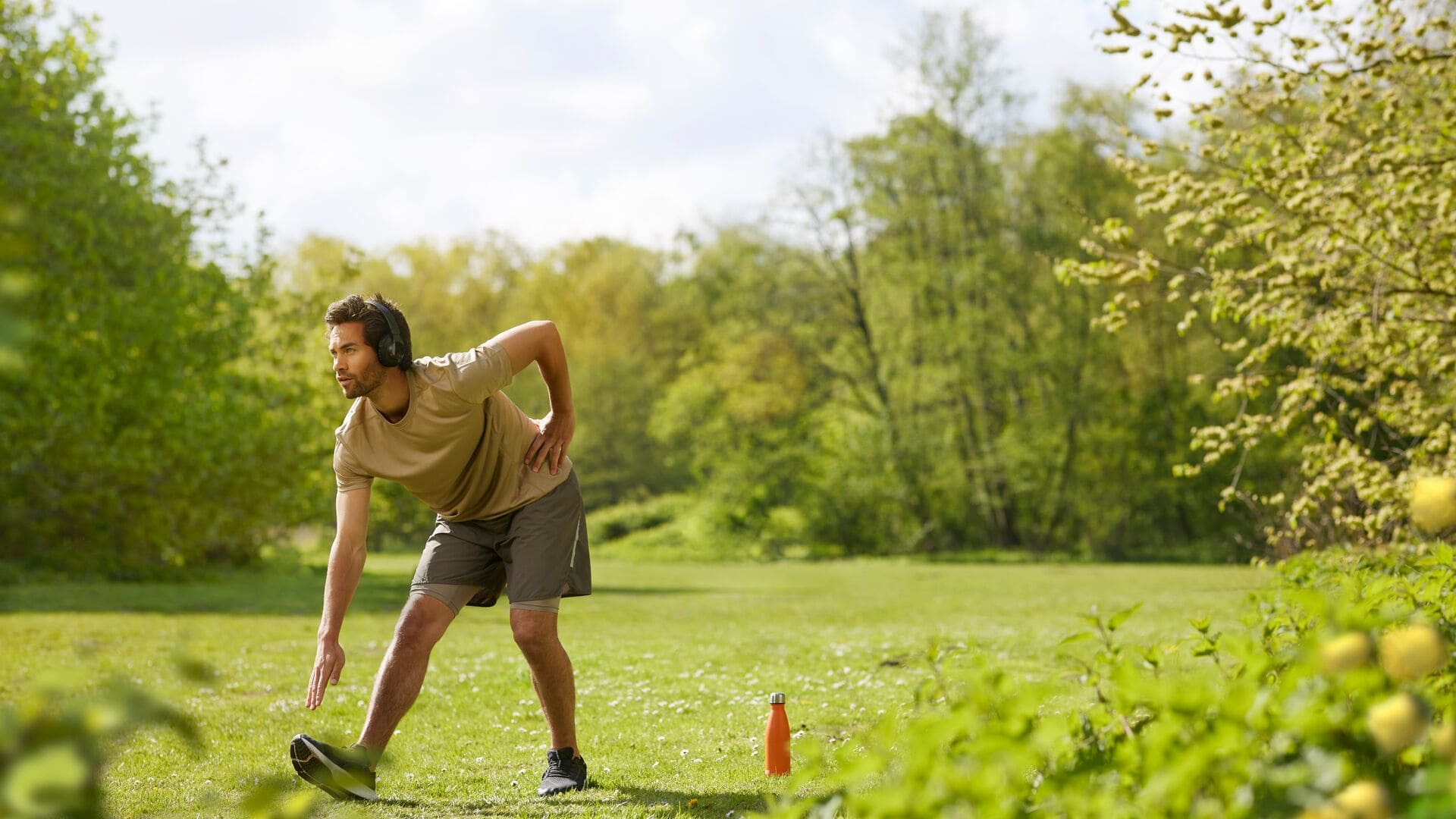 A man in a beige t-shirt and shorts stretches outdoors on the grass, wearing headphones, with an orange water bottle placed nearby.