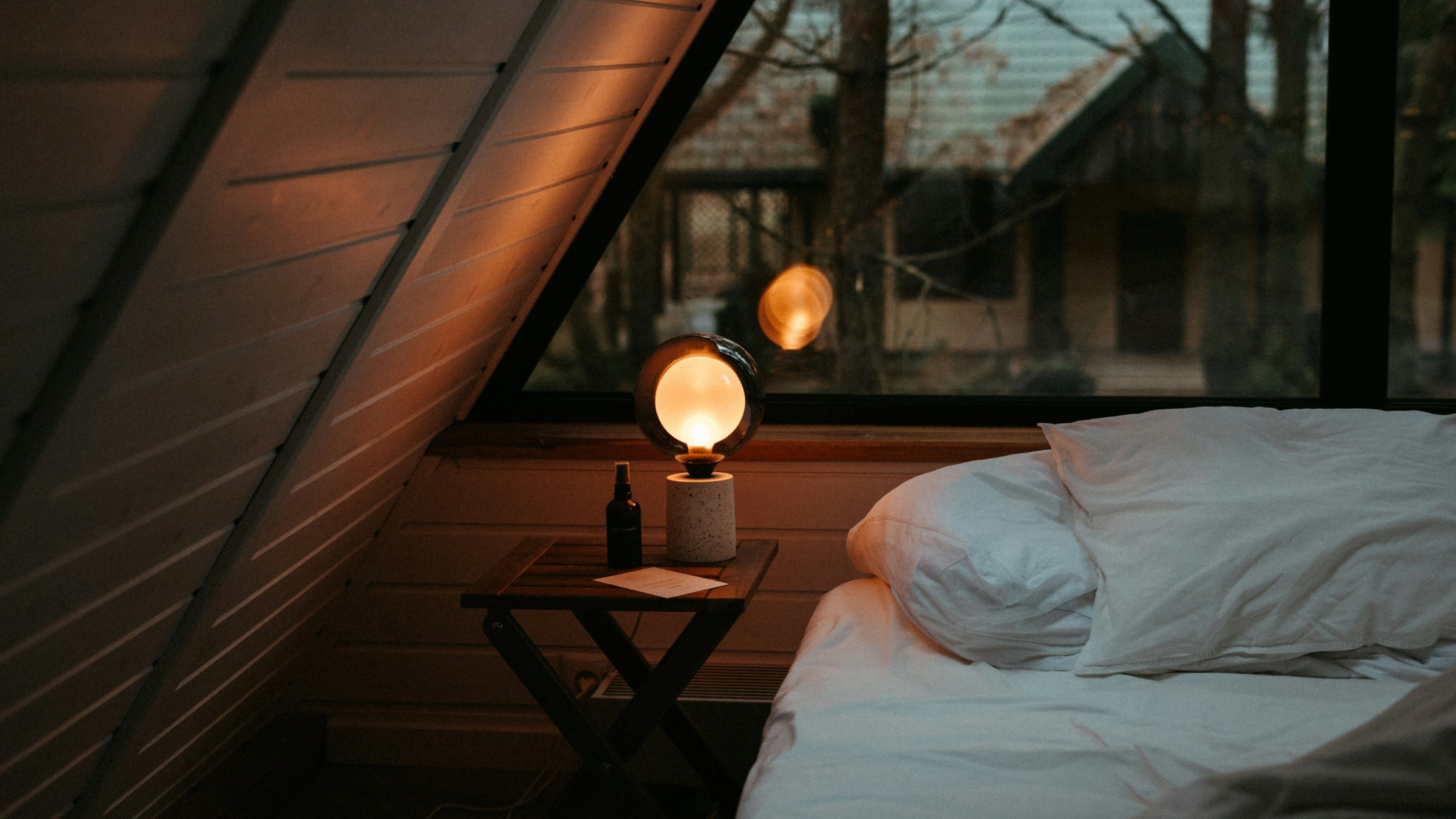 A cozy attic bedroom with white bedding and a glowing lamp on a wooden nightstand, creating a warm and calm evening atmosphere.