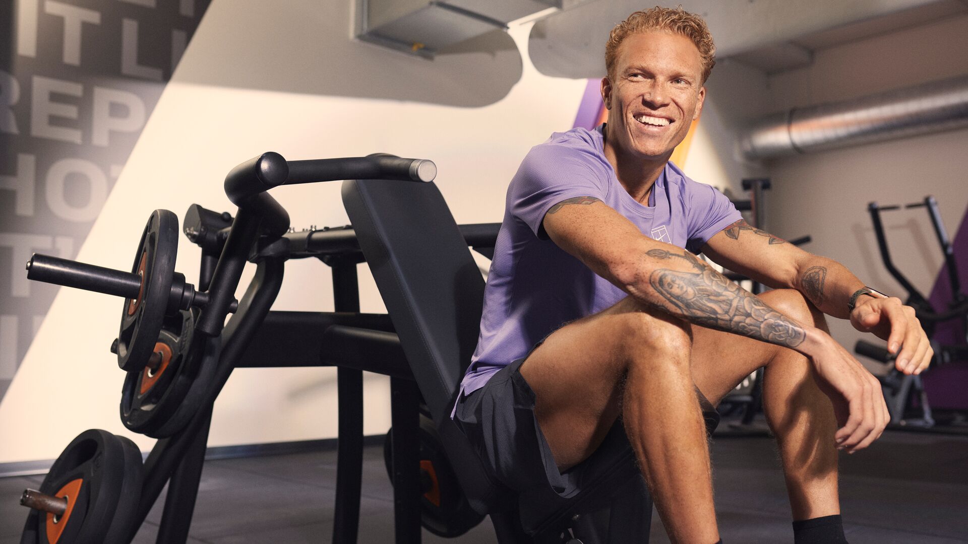 Man sitting on a strength machine in a gym, smiling after a workout, wearing a purple T-shirt and black shorts.