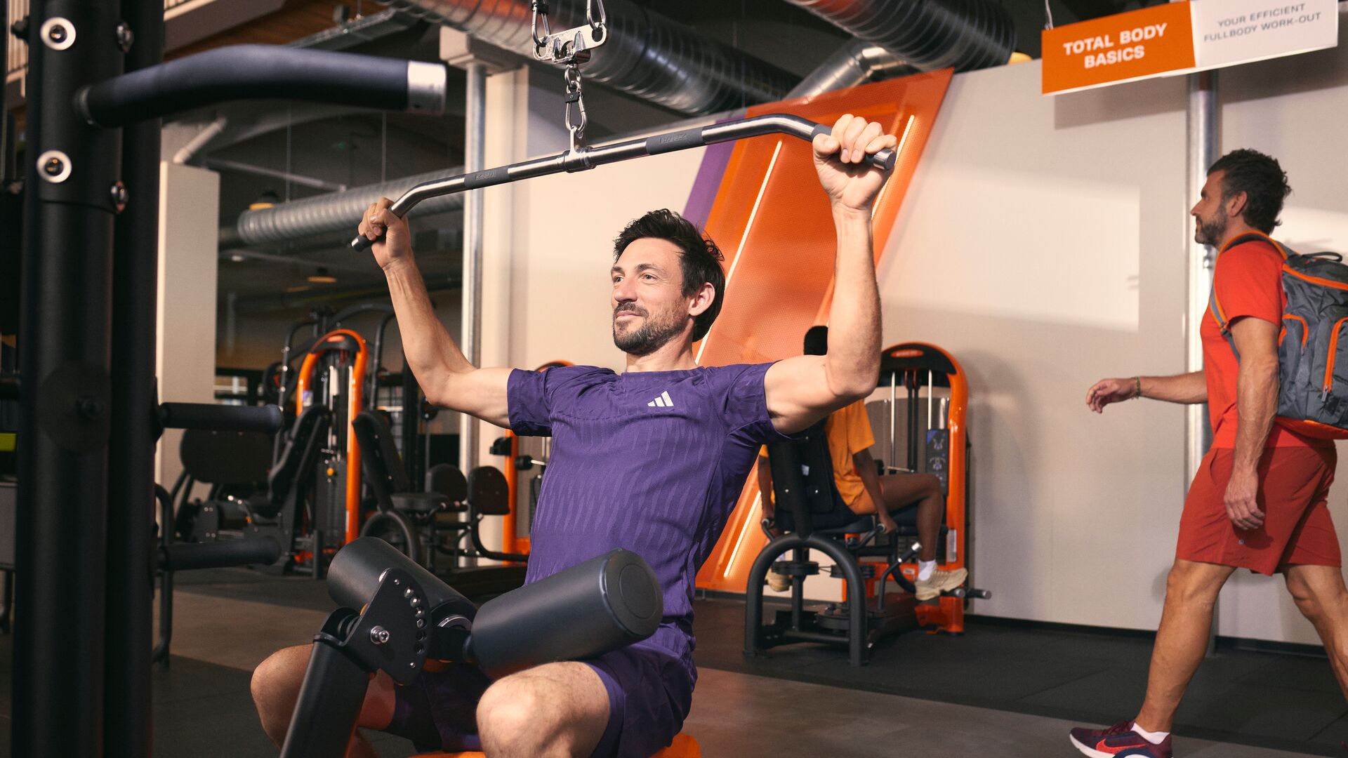 Man performing a lat pulldown exercise on a fitness machine in a modern Basic-Fit gym.