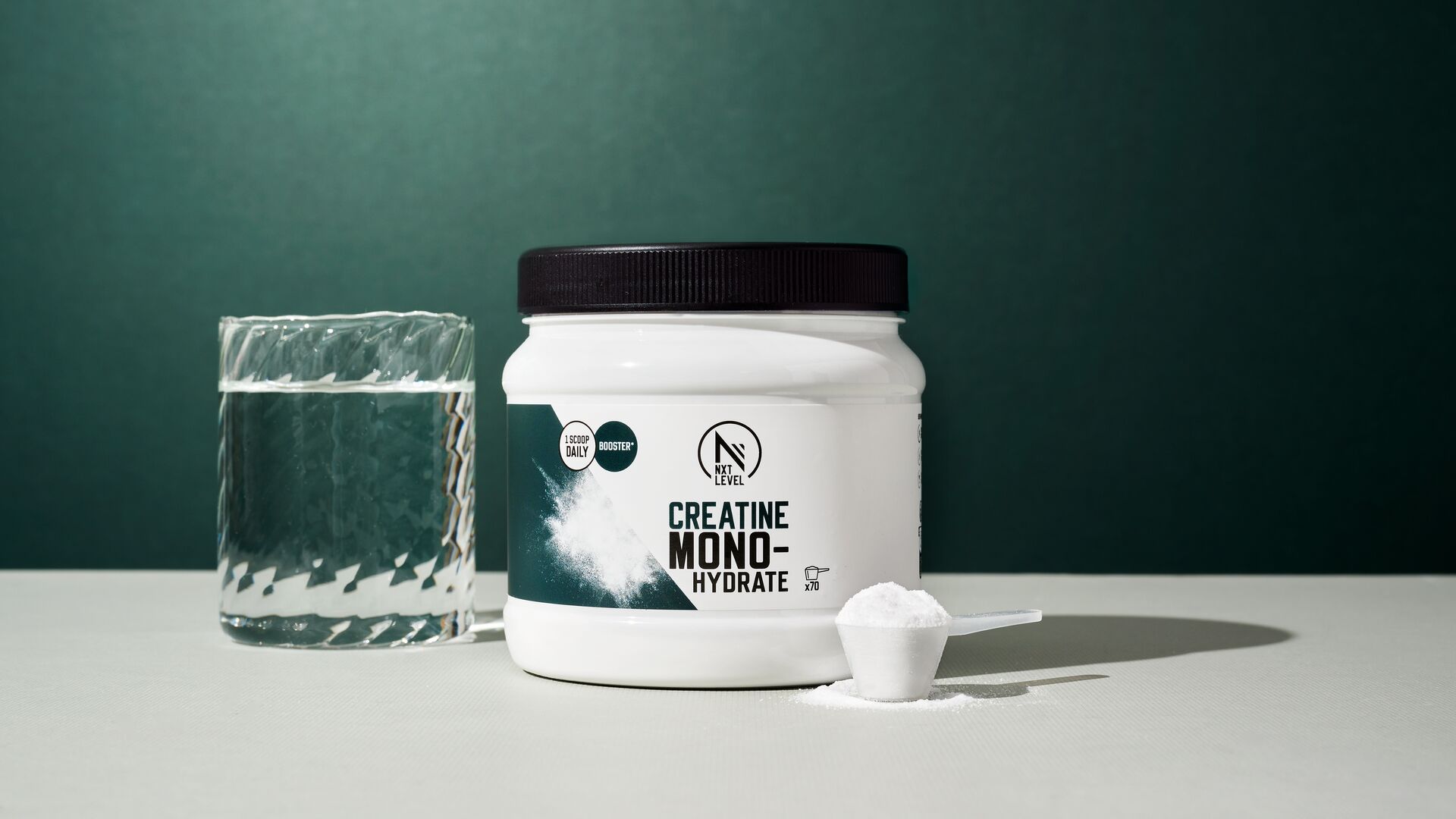 A container of creatine monohydrate sits on a table next to a glass of water and a scoop filled with powder, photographed against a dark green background.