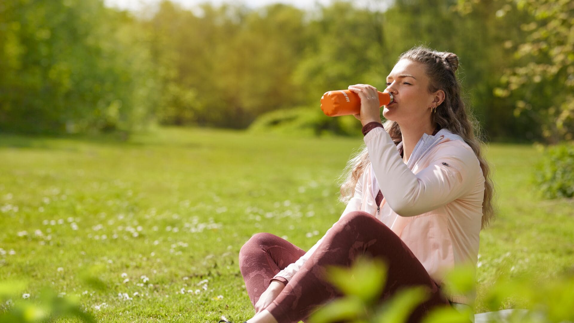 A woman enjoys a break in the park, sitting on the grass and drinking from her orange Basic-Fit bottle.