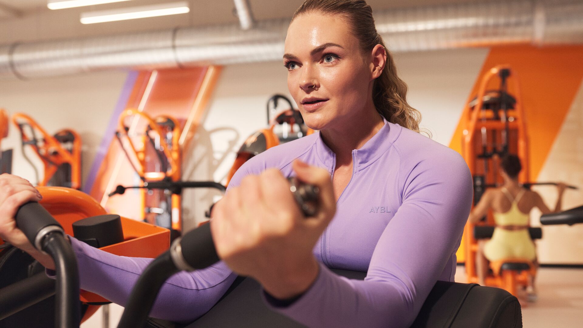 Person using a bicep curl machine in a gym, focusing on arm engagement while seated, with orange strength machines visible behind.